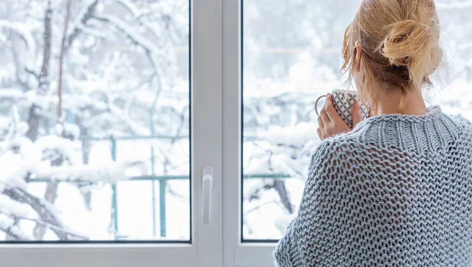 Woman in gray sweater drinking coffee by window with snow outside